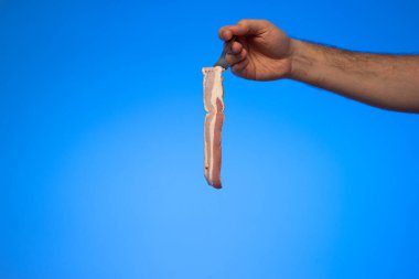 Mavi arka planda tek dilim çiğ domuz pastırması, beyaz erkek eli tarafından elde tutulan bir çatal üzerinde..