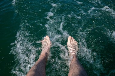 Kafkas erkek bacakları ve ayakları berrak, turkuaz suya batmış, küçük kiralık sürat teknesinin kıç güvertesinde sıçrama ve dalgalanmalar yaratıyor. POV perspektifi, aşağı bakıyor