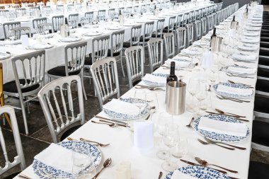 Sandalyeler, masa örtüleri, tabaklar, çatal bıçak takımı ve bardaklarla dolu uzun masalar. Geniş açı, iç mekan, yukarıdan aşağı, insan yok.