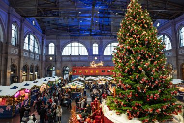 Zürih HB, İsviçre, 26 Kasım 2025. Büyük, uzun boylu, Lindt ana istasyon binasındaki Noel ağacını süsledi ve insanlar birçok yiyecek ve içecek tezgahını ziyaret etti. Geniş açı görünümü