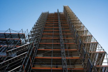 İnşaat veya yenileme sırasında yüksek binaları kapsayan geniş bir yapı iskelesinin alçak açılı görüntüsü. Karmaşık metal yapısı parlak mavi bir gökyüzüne doğru yükseliyor.