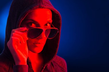 Kapüşonlu ve güneş gözlüklü genç bir kadının portresi, koyu mavi bir arka planda, kırmızı ışıkla aydınlatılmış..