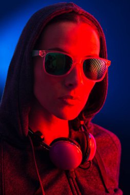 Kapüşonlu, güneş gözlüklü ve kulaklıklı genç bir kadının portresi, koyu mavi bir arka planda, kırmızı ışıkla aydınlatılmış..