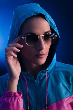 Kapüşonlu ve koyu mavi arka planda güneş gözlüğü takan genç bir kadının portresi..