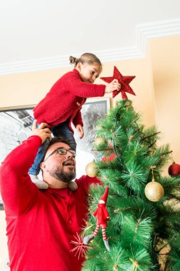 Kırmızı kazak giyen bir adam ve bir kız yıldızı Noel ağacının tepesine asıyor..