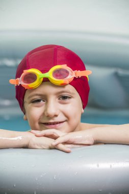 Kırmızı yüzme şapkalı ve su gözlüklü bir çocuk, küçük lastik bir havuzda, iki su balonu gösteriyor. Yaz, su oyunları için ideal bir zamandır..