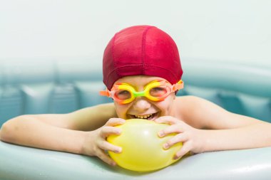Kırmızı yüzme şapkalı gülümseyen bir çocuğun portresi, küçük lastik bir havuzda, sarı bir su balonuyla oynuyor. Yaz, su oyunları için ideal bir zamandır..