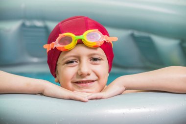 Kırmızı yüzme şapkalı ve su gözlüklü bir çocuk, küçük lastik bir havuzda, iki su balonu gösteriyor. Yaz, su oyunları için ideal bir zamandır..