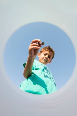 Çocuk çöp kutusunun içine bir yığın yerleştiriyor, arka planda mavi bir gökyüzü ile çöp kutusunun içinden görülüyor. Çevre değerlerinde geri dönüşüm ve eğitim kavramı.