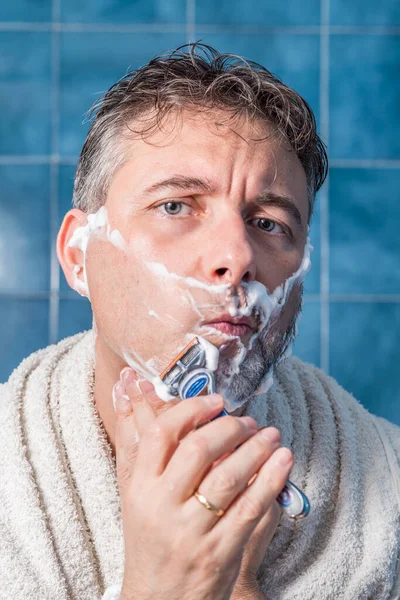 中年男子 肩上披着白毛巾 剃须刀刮胡子 背景是蓝色瓷砖 男子个人护理的概念 — 图库照片