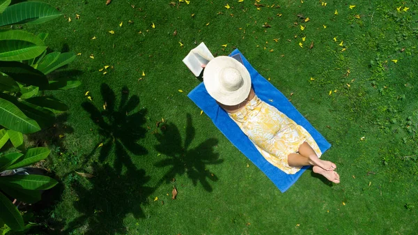 Молода жінка в одязі читає книгу, що лежить на зеленій траві в тропічному саду в сонячний день . — стокове фото