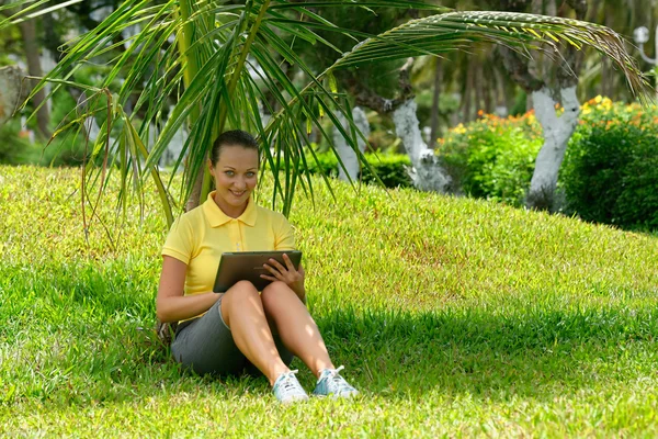 Jovem mulher usando tablet deitado ao ar livre na grama, sorrindo . — Fotografia de Stock