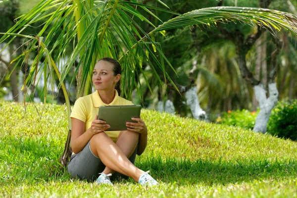 Jonge vrouw op het gazon onder een palmboom met haar tablet comput — Stockfoto