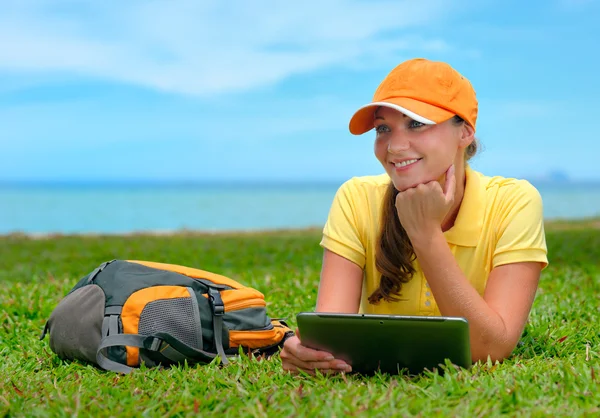 バックパックとタブレット c と芝生に横になっている若い女性の笑みを浮かべてください。 — ストック写真