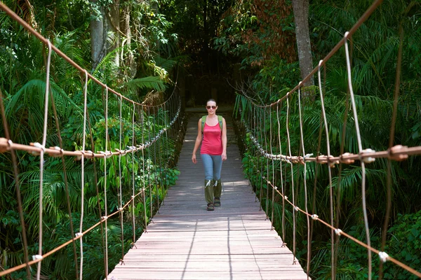 Mujer feliz excursionista cruzando puente colgante a la luz del sol . — Foto de Stock