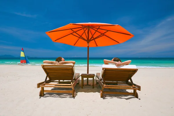 Casal em uma praia tropical em cadeiras de convés sob um guarda-chuva vermelho — Fotografia de Stock