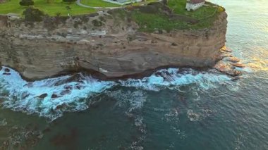 Los Angeles, Kaliforniya 'daki Point Fermin Deniz Feneri' nin renkli gökyüzü, engebeli uçurumlar ve kıyıya vuran dalgalarla birlikte sinematik hava yörüngesi görüntüsü.