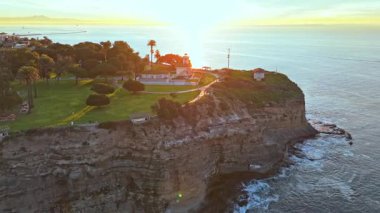 Los Angeles, Kaliforniya 'daki Point Fermin Deniz Feneri' nin renkli gökyüzü, engebeli uçurumlar ve sahil boyunca dalgaların çarptığı hava yörüngesi görüntüsü.