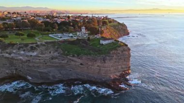 Los Angeles, Kaliforniya 'daki Point Fermin Deniz Feneri' nin renkli gökyüzü, engebeli uçurumlar ve sahil boyunca dalgaların çarptığı hava yörüngesi görüntüsü.