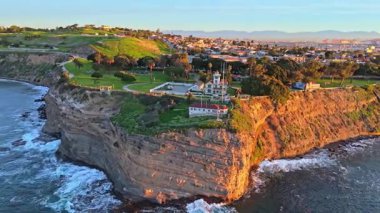 Los Angeles, Kaliforniya 'daki Point Fermin Deniz Feneri' nin renkli gökyüzü, engebeli uçurumlar ve sahil boyunca dalgaların çarptığı hava yörüngesi görüntüsü.