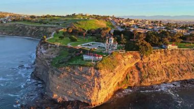 Los Angeles, Kaliforniya 'daki Point Fermin Deniz Feneri' nin renkli gökyüzü, engebeli uçurumlar ve sahil boyunca dalgaların çarptığı hava yörüngesi görüntüsü.