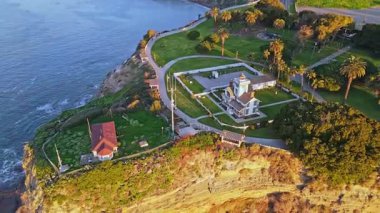 Los Angeles, Kaliforniya 'daki Point Fermin Deniz Feneri' nin renkli gökyüzü, engebeli uçurumlar ve kıyıya vuran dalgalarla çekilen sinematik görüntüsü.