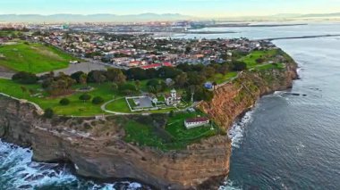 Los Angeles, Kaliforniya 'daki Point Fermin Deniz Feneri' nin renkli gökyüzü, engebeli uçurumlar ve kıyıya vuran dalgalarla altın saatinde çekilmiş yüksek hava sarmalı görüntüsü.