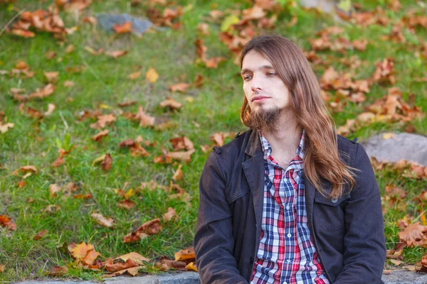 Man långt hår sitter på bänken i höst park — Stockfoto