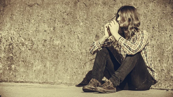 Uomo dai capelli lunghi seduto solo triste sul muro grunge — Foto Stock