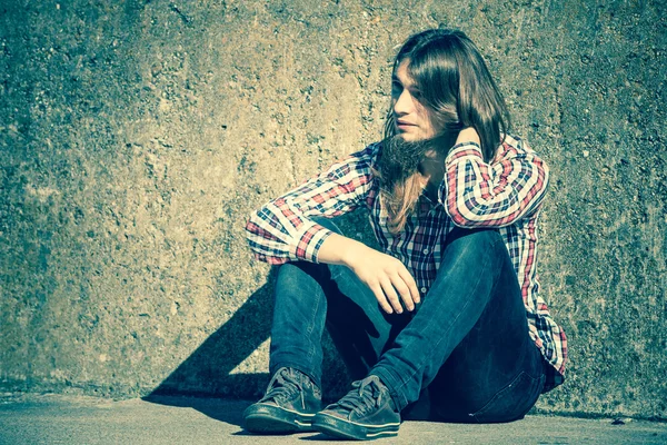 Hombre de pelo largo sentado solo triste en la pared grunge — Foto de Stock