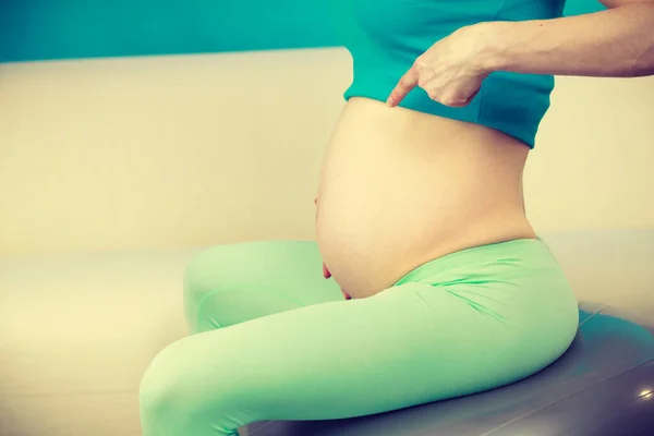 怀孕妇女舒适地坐在舒适的瑞士球在家里 手指着她的腹部 健康和健康生活方式概念 — 图库照片