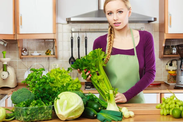 Woman in kitchen with many green leafy vegetables, fresh produce on table. Young blonde female adding to her diet foods high in chlorophyll. Healthy eating, vegan food, fiber for weight loss.