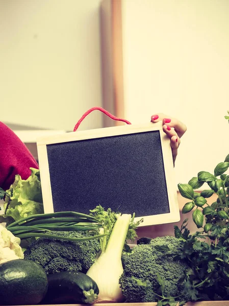 キッチンの若い女性は 健康的で菜食主義者の何かを調理するために多くの緑の野菜を持っています コピースペースのための空白の黒い板を保持 — ストック写真