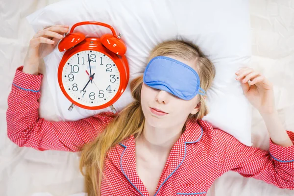 Sleeping young woman wearing cute pink pajamas holding big red old fashioned clock showing sleep time.