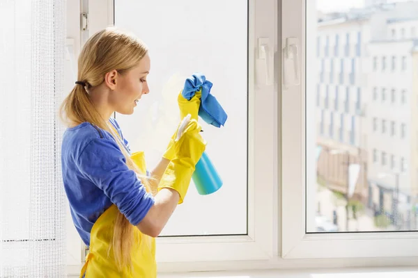 穿着手套和围裙的年轻女子在家里用抹布和喷雾清洁剂洗窗玻璃 蓝色的黄色 清洗概念 — 图库照片