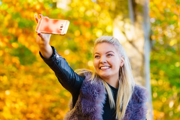 秋の公園を歩く おしゃれな衣装の人工毛皮のコートを着た金髪の魅力的な女性は スマートフォンで自撮り写真を撮る 幸せな正の女性が楽しい — ストック写真