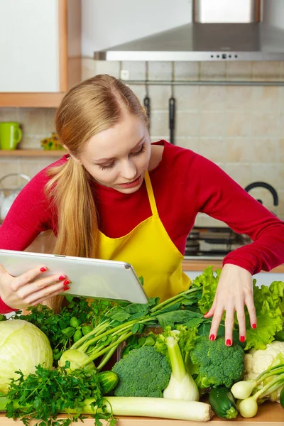 台所の若い女性はテーブルの上に多くの緑の野菜を持っています 何かを調理し インターネットでレシピを検索することについて考えるタブレットを保持 — ストック写真