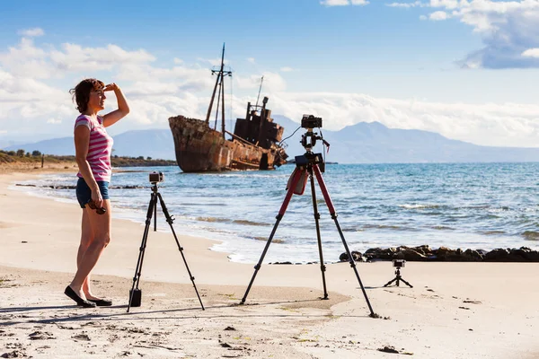 錆びた難破船と海岸線からの写真フィルムビデオを撮る三脚のプロのカメラを持つ観光女性写真家 — ストック写真