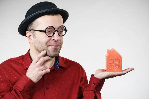 面白い帽子と眼鏡をかぶった大人の男が小さな赤い家モデルを保持しています 不動産屋になろうと思ってる 住宅所有権の概念 — ストック写真
