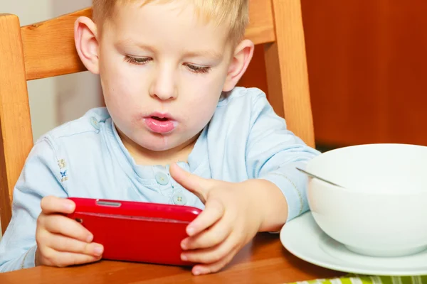 携帯電話で遊んで食べてよだれをたらしている少年朝食します。 — ストック写真