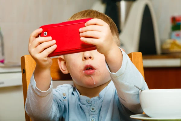Chlapec slintání stravování snídaně, hraní s mobilním telefonem — Stock fotografie