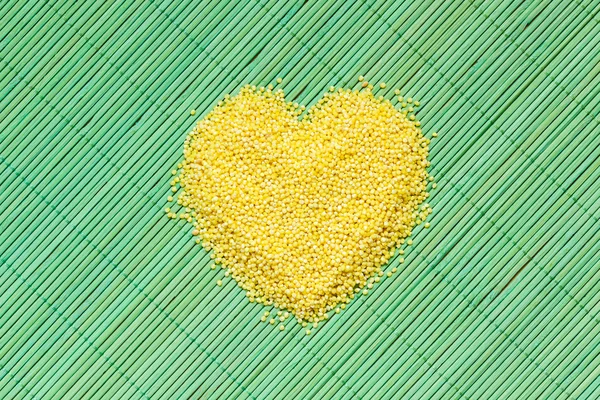 Millet groats heart shaped on green mat surface. — Stock Photo, Image