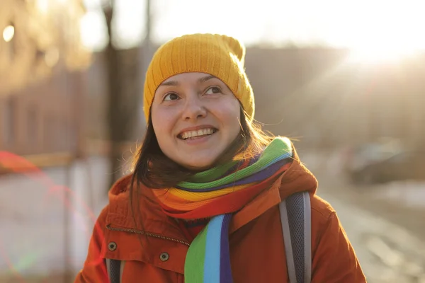 Mignon fille heureuse portant casquette jaune et écharpe arc-en-ciel souriant à la caméra avec soleil briller — Photo