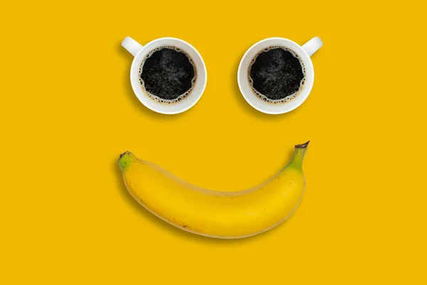 Happy morning concept. Two coffee cups and a banana shot on an bright yellow background. The composition of the objects looks like a smiley face.