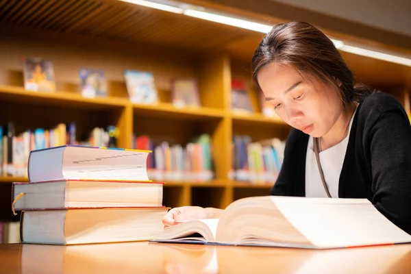 A lovely Asian woman reads a book in the university library to relax and educate herself. A large book on a wooden table