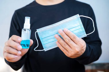 Alkol spreyi ve maskeyle el dezenfektanı tutan adam. COVID-19 salgını sırasında olması gereken şeyler.