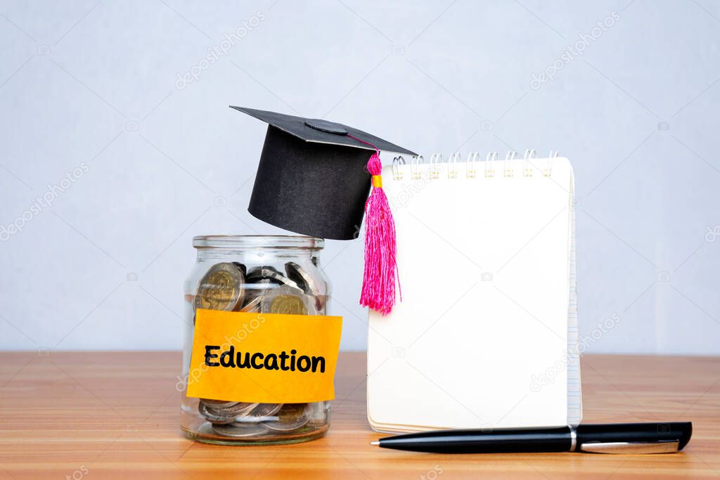 Graduation hat and glass jar with money, pen and a blank notebook for education on wooden table