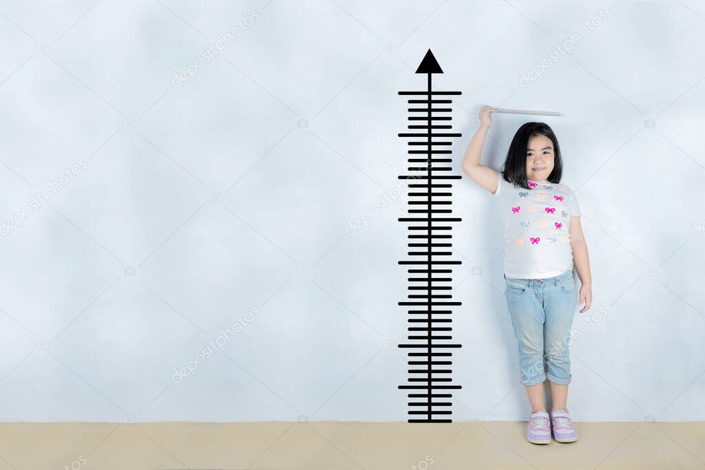 A little Asian girl measures the height near the white wall.