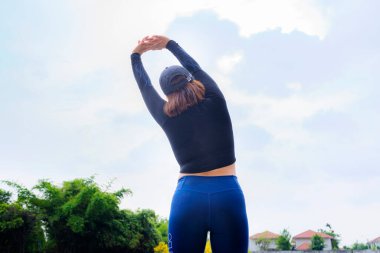 Spor kıyafetleri içinde genç ve güzel bir Asyalı kadın sağlıklı bir yaşam tarzı elde etmek için sabah spordan önce parkta esneme hareketleri yapıyor. Sağlıklı genç bir kadın dışarıda ısınıyor..