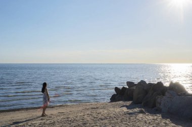 Beyaz elbiseli genç sarışın kadın kumların üzerinde tek başına duruyor ve sakin denize ve açık mavi gökyüzüne bakıyor..